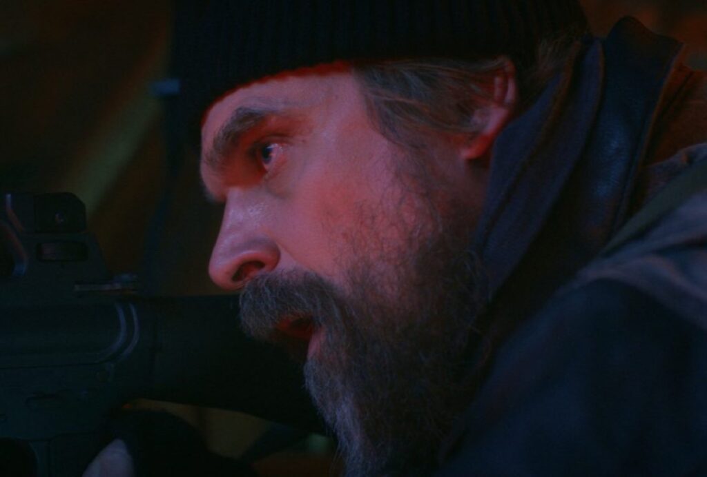 bearded middle aged man in ski cap pointing a rifle.