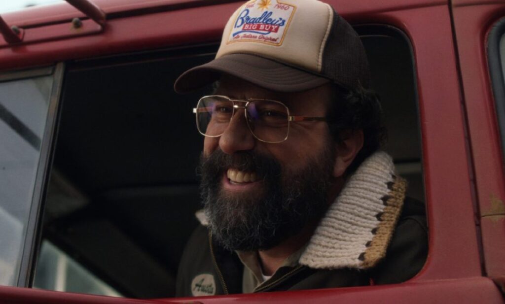 bearded man with glasses and a baseball cap smiling from the cab of a red truck.