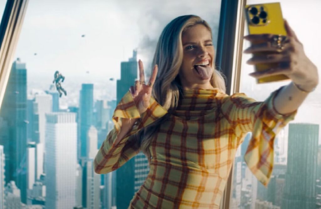 blond woman in checked yellow dress taking a selfie while sticking her tongue out and making a V for victory sign with her fingers.