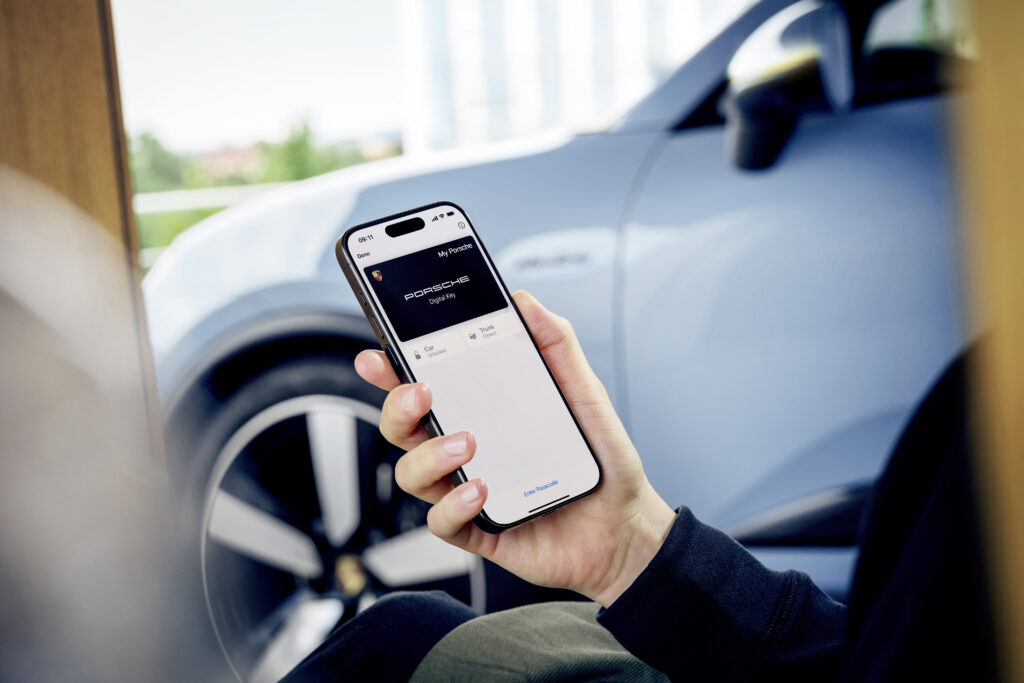 A man holds a smartphone with a Porsche digital key on it, as a Macan charges nearby.