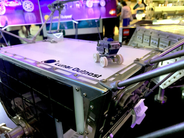 a miniature white and blue model of a moon rover made out of building brick toys atop a full-size mockup of the same rover