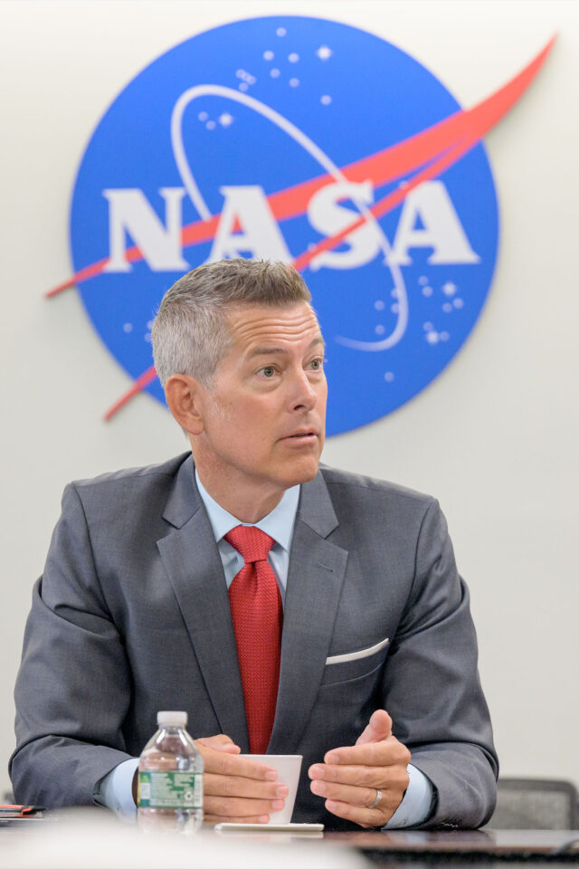 a man with gray hair and pale complexion wears a gray suit and red tie while sitting at a table under a red, white and blue NASA logo on the wall behind him