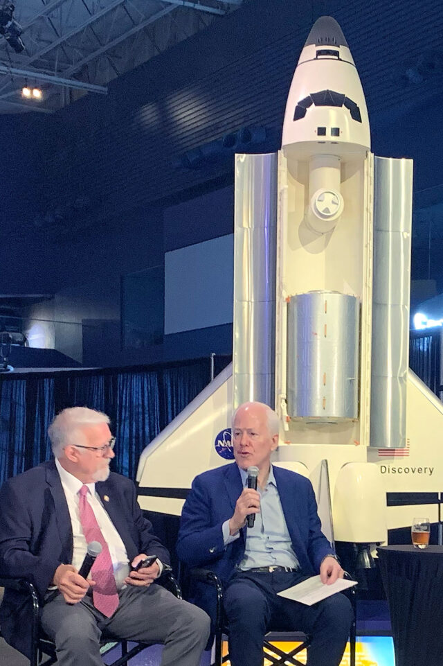 two men in business suits sit front of a large model of a space shuttle