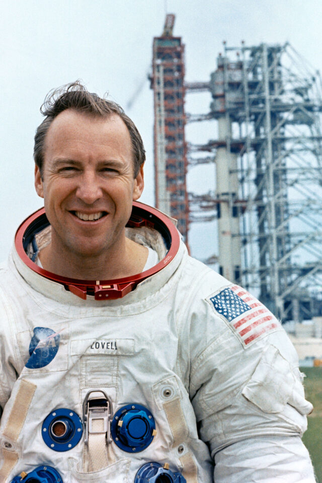 a man in a white spacesuit stands in front of a launch pad where a rocket is being prepared for his mission