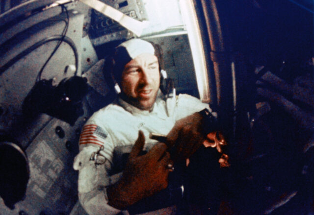 a man is seen wearing a white coveralls and brown head cap inside a spacecraft