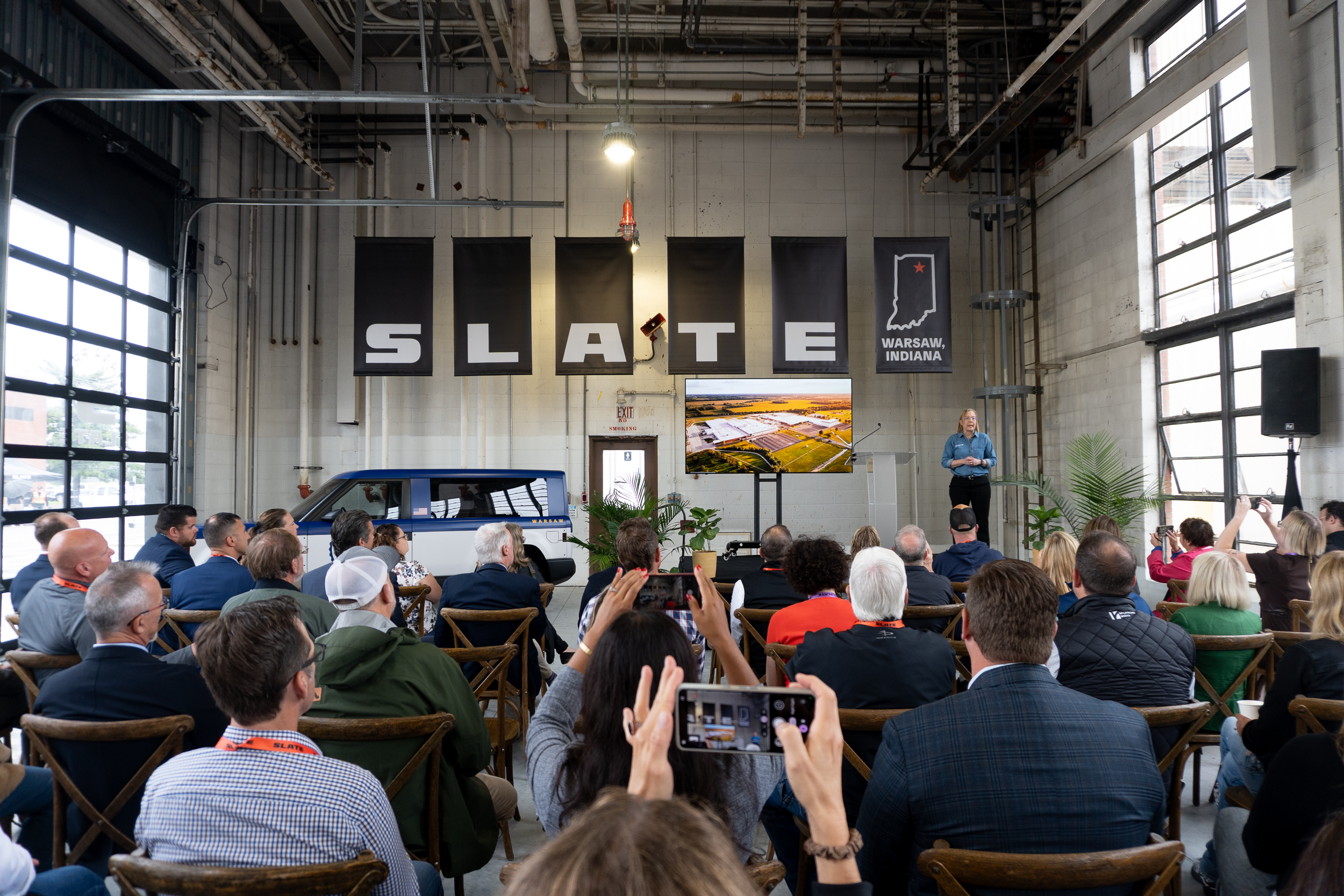 A woman stands at a podium in front of an audience. To her right is a small pickup truck.
