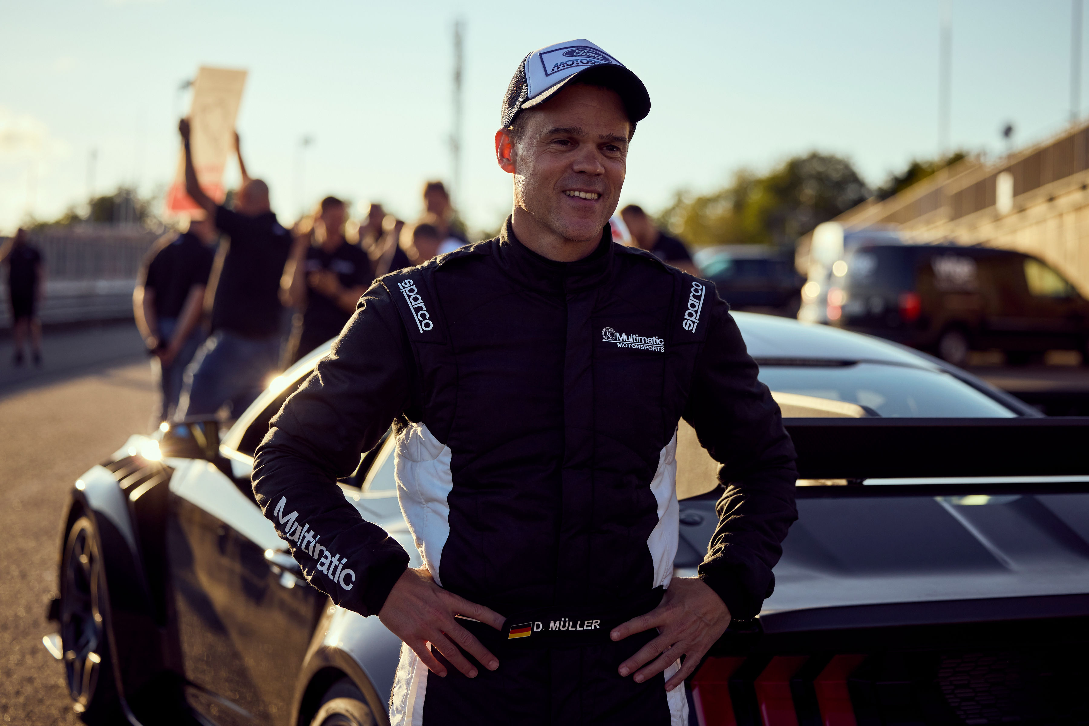 A racing driver stands in front his car as mechanics and engineers celebrate in the background.