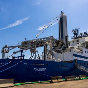 Image of a large blue ship with specialized hardware and cranes on its deck, tied up to a brown dock.