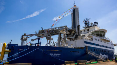 Image of a large blue ship with specialized hardware and cranes on its deck, tied up to a brown dock.