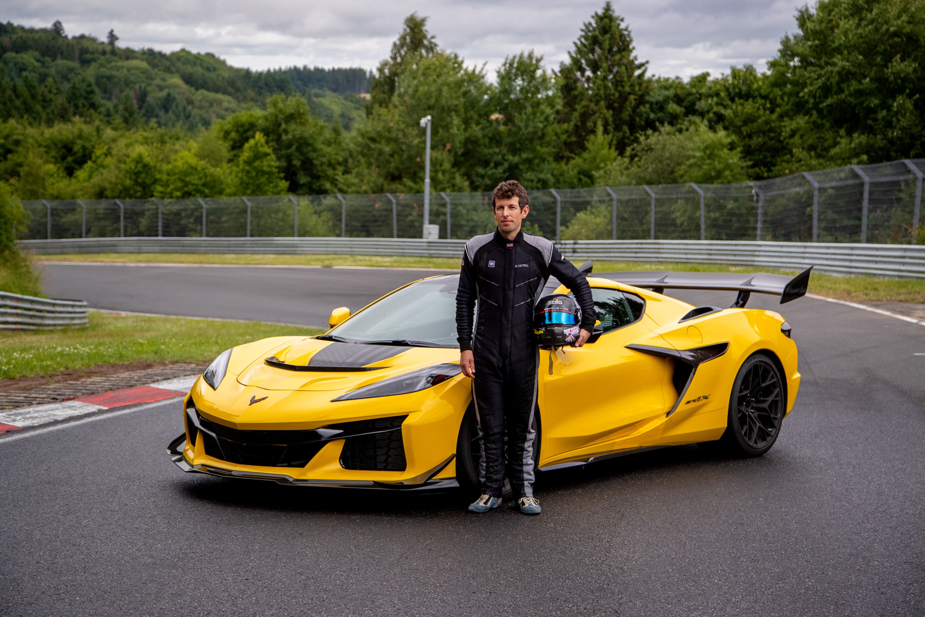 An engineer in a fire suit stands next to a yellow Corvette, parked on the Nurburgring.
