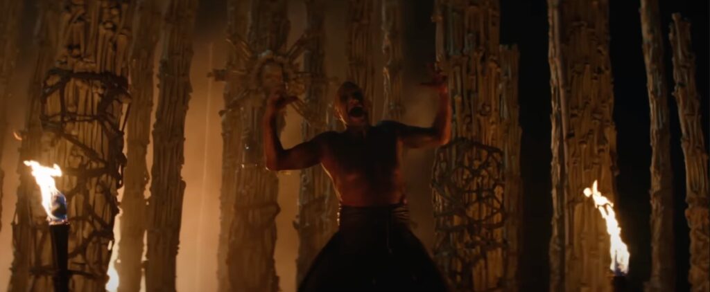 Hombre de mediana edad de Chested Bare de pie de pie en el centro de un templo de los huesos, chillando hacia arriba con los brazos elevados desafiantes.