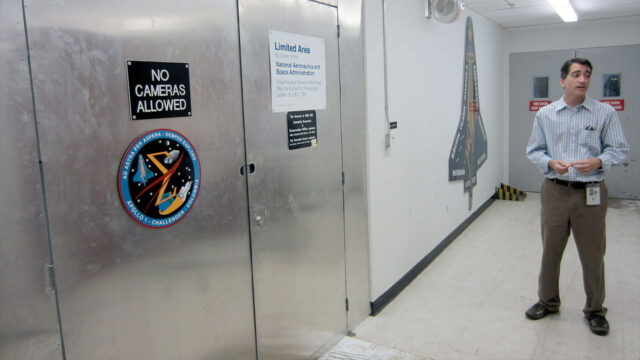 a man in a button down shirt and slacks stands outside the steel door entrance to a limited access room.