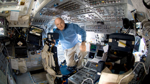 a man in a blue shirt and khaki slacks is seen surrounded by switches and displays aboard a spacecraft