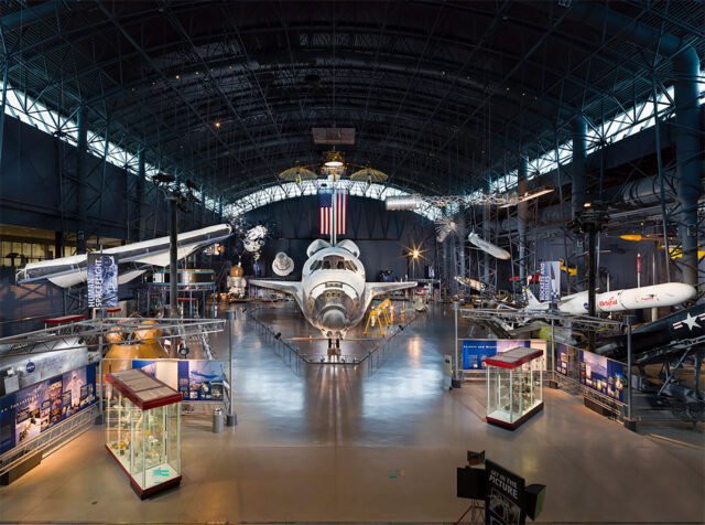 An overhead view of museum exhibits, including a space shuttle, inside a large museum hangar