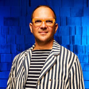 Balding man wearing striped shirt and jacket standing against blue background with hands in pockets.
