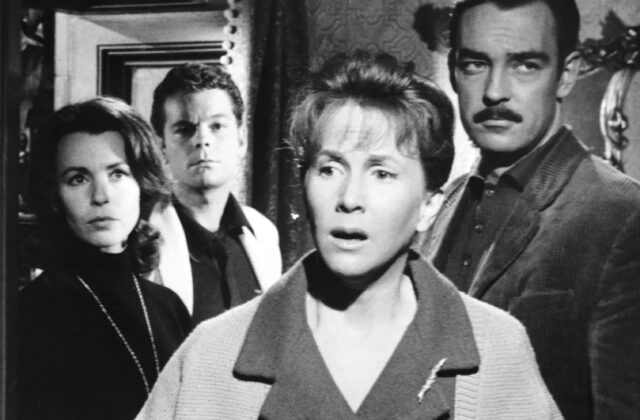 B&W still of group of people in 1960s clothing standing in drawing room of a haunted house