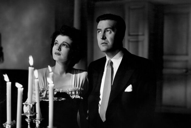 B&W image of man and woman in 1940s evening wear holding a candle and looking up a flight of stairs