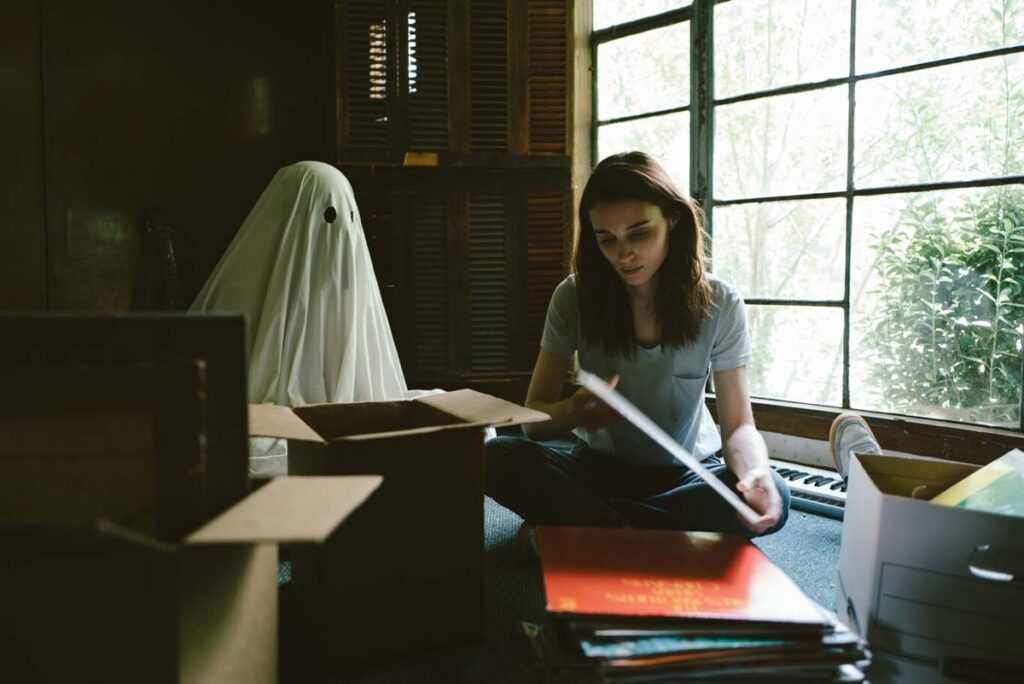 young woman seated at a desk with a small figure draped in a sheet wth eye holes cut out standing beside her