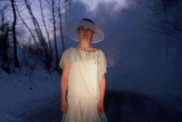 young woman with dark haired bob wearing a 1920s white dress and hat, standing in a road illuminated by headlights on a snowy night