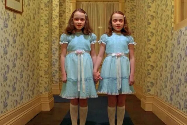 twin girls in matching light blue dresses and white knee socks standing in a hallway with yellow flowered wallpaper