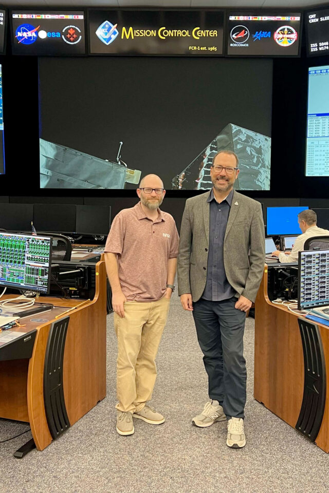 two men pose together for a photo in a mission control room