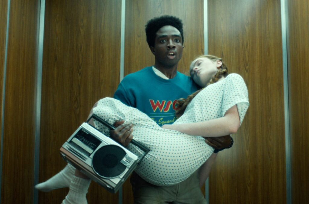 Young black man carrying a young girl in hospital PJs in an elevator, carrying a boom box in one hand.