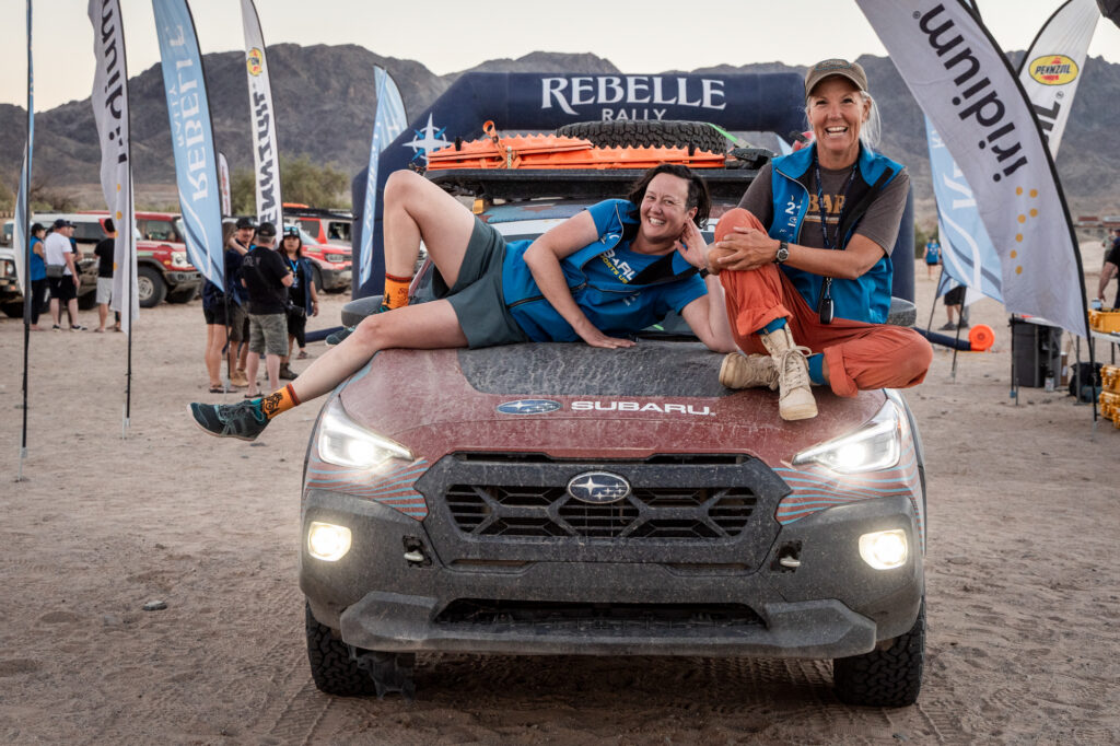 two women rally drivers sit on the hood of their car at the end of the race