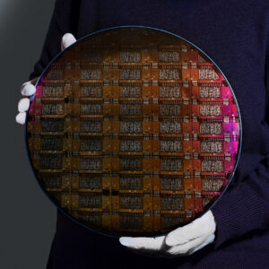 A person in a dark blue lab coat with white gloves holds a large disk with many circuits printed on it.