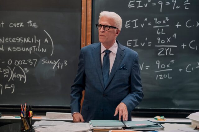 White haired older man in a nice blue suit and tie standing in front of a blackboard filled with equations in a college classroom