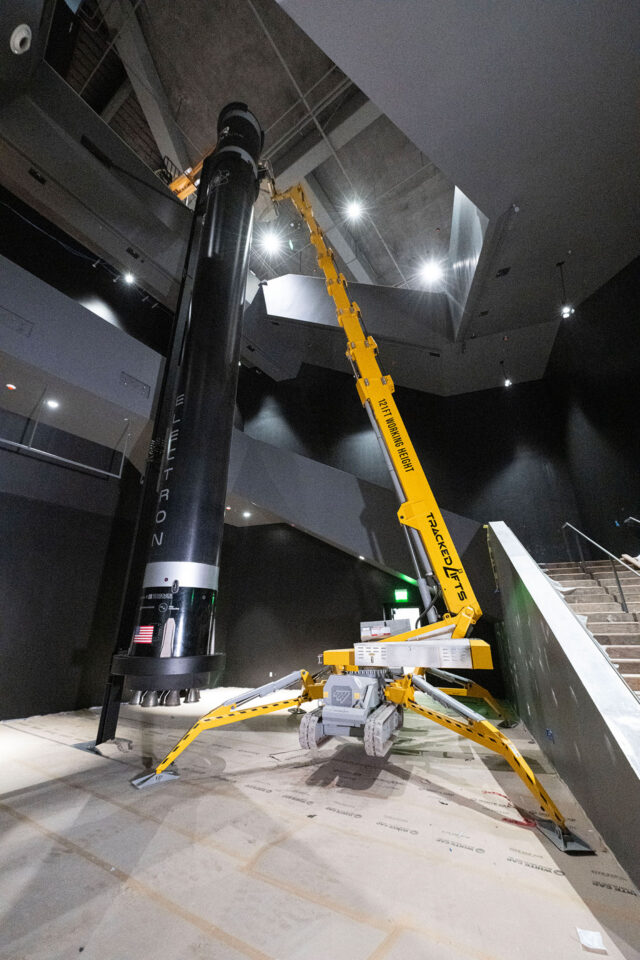 a large yellow crane is used to lift a long, black cylindrical artifact into place inside a museum