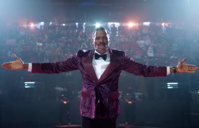 black man in colorful purple suit with arms outstretched in front of a camera with huge crowd behind him