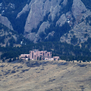 Image of a dry, grassy hill at the foothills of rocky, tree covered mountains. A large cluster of brown buildings sits at the peak of the hill.