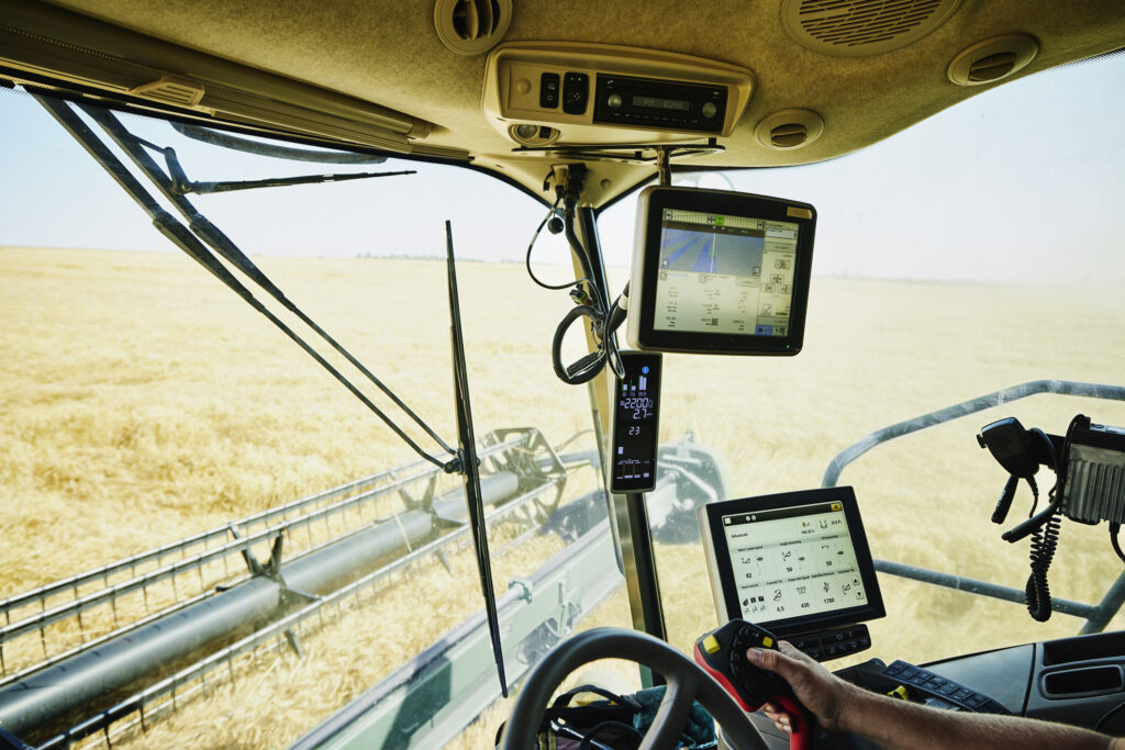 Image of the inside of the cabin of a large farming machine moving through a field of wheat. Screens track its current location and where it has been.