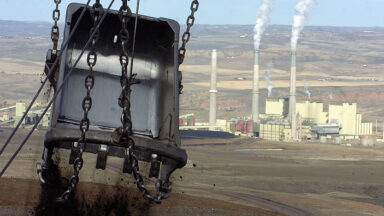 Image of a large metallic shovel suspended from chains in the foreground, with a coal generating plant in the background.