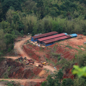 A hillside mining operation with three long buildings on top of the hill. A dirt road with construction equipment leads to the buildings at the top.