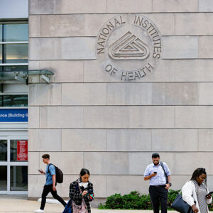 People standing near a bus stop in front of a large stone building facade with the NIH logo etched into the stone.