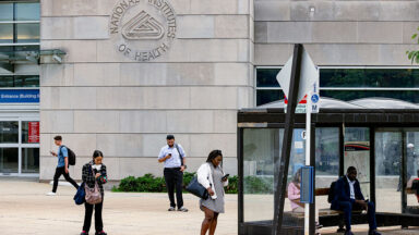 People standing near a bus stop in front of a large stone building facade with the NIH logo etched into the stone.