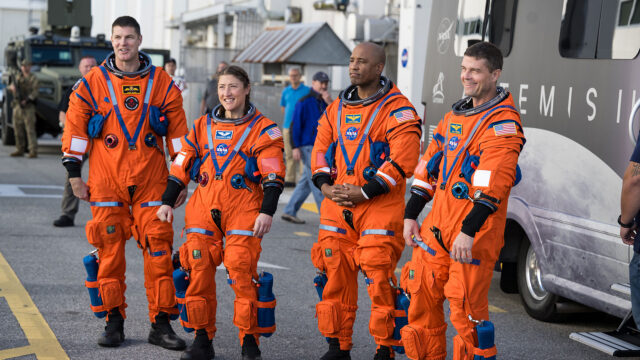 Three men and a woman wearing bright orange pressure suits pose for a photo next to a motor coach.