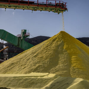 A massive, industrial-scale pile of yellow sulfur, with a conveyor belt dumping more on.