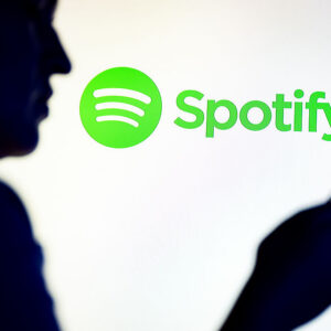In this photo illustration, a silhouetted individual is seen holding a mobile phone with a Spotify logo displayed in the background.