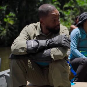 actor will smith in khakis seated with arms folded on is knees in a canoe rowing down a tropical river