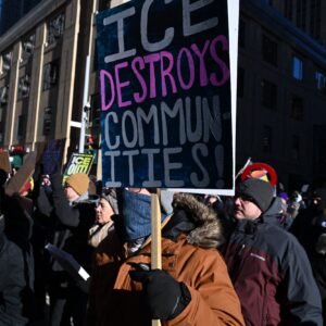 People wearing winter coats, hats, and gloves take part in a march against US Immigration and Customs Enforcement (ICE) in Minneapolis, Minnesota. One person holds a sign that says, "ICE destroys communities!"