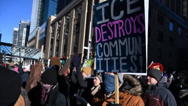 People wearing winter coats, hats, and gloves take part in a march against US Immigration and Customs Enforcement (ICE) in Minneapolis, Minnesota. One person holds a sign that says, "ICE destroys communities!"