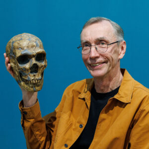 Image of a man with glasses and a mustard colored jacket holding up a skull.