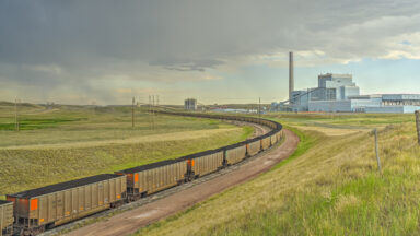 A grass covered prairie with rail line curving around to the left. The rails are filled with train cars carrying coal, and a large facility with a smokestack sits on the horizon.