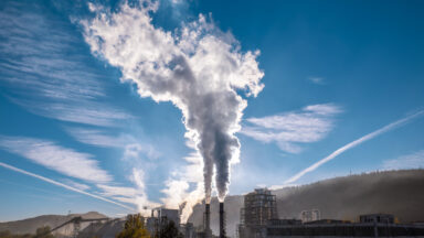 Image of a blue sky with a hazy industrial facility in the lower foreground. Two large smokestacks are releasing a cloud of material into the air.