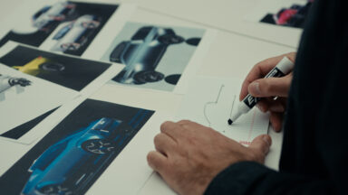 a man's hands sketch a pickup truck outline, with pictures of other trucks on his bench