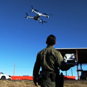 A US Border Patrol agent holds a controller while a drone flies overhead.