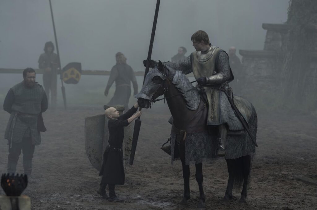 small bald boy handing a lance to a knight on a horse