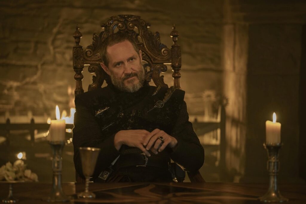 dark haired man with graying beard seated at a table with a chalice of wine at his elbow